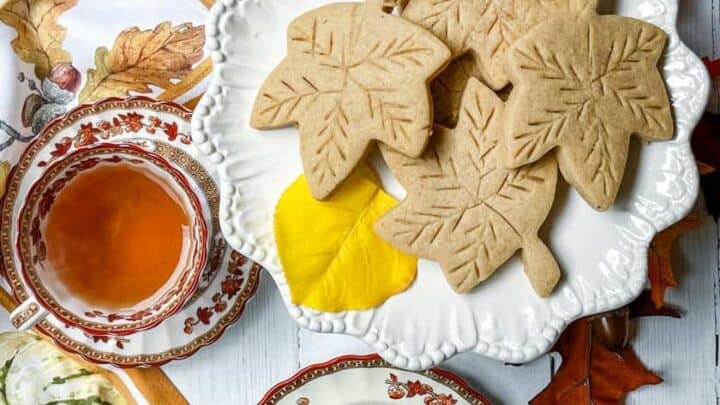 A white plate with leaf-shaped spice sugar cookies sits on a table surrounded by autumn leaves. Two teacups filled with tea and matching saucers are nearby, along with a decorative napkin featuring fall motifs.