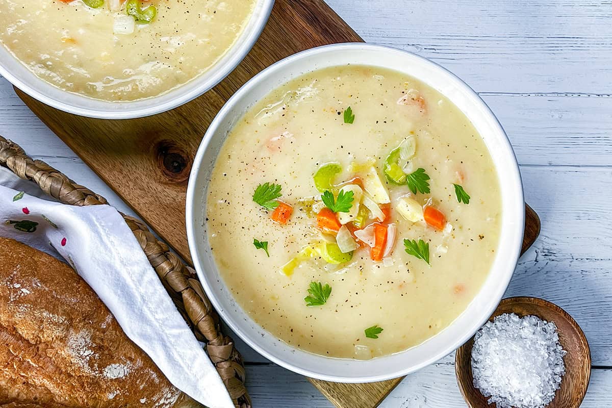 A bowl of creamy potato leek soup garnished with herbs and chopped vegetables, served on a wooden board with bread and a small dish of salt nearby.
