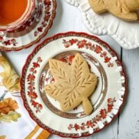 A maple leaf-shaped spice cookie on a decorative plate sits on a white wooden table. A cup of tea accompanies it, alongside a floral napkin. Another plate with sugar cookies is partially visible, hinting at an inviting spread.