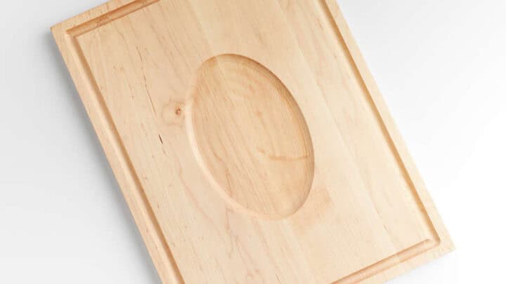 A rectangular wooden cutting board with a groove near the edge and an oval-shaped depression in the center, this Thanksgiving kitchen essential tool is photographed against a white background.