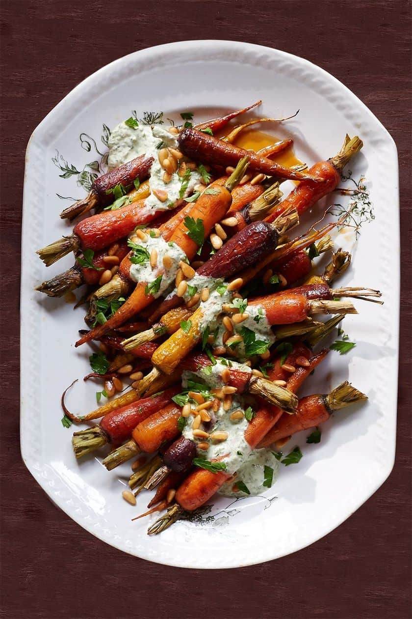 Roasted baby carrots with green tops, garnished with a creamy white sauce, chopped herbs, and pine nuts on a white oval platter-a festive addition to your Christmas side dishes.