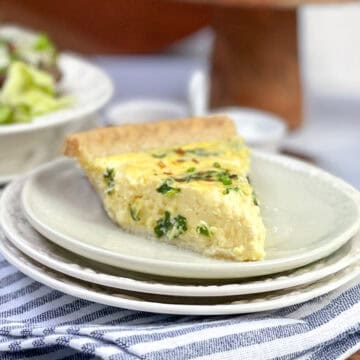 Slice of spinach quiche on a white plate, ready for serving, with a green salad in the background.