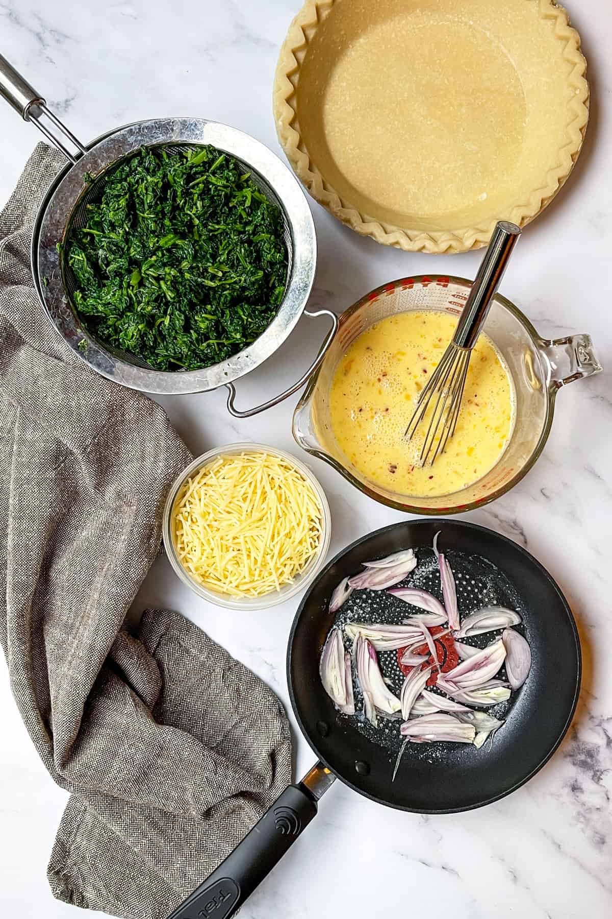 Spinach Quiche ingredients on a marble surface including eggs, shredded cheese, spinach and saut&eacute;ed shallots.