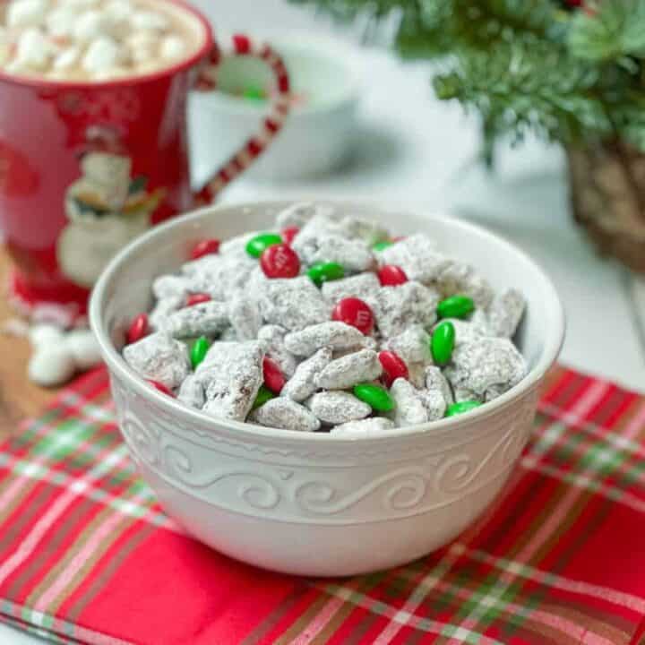 Christmas Muddy Buddies in a White Bowl with Hot Chocolate in the background