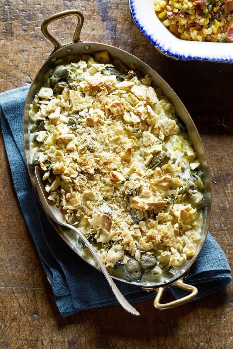Oval baking dish with a creamy casserole topped with crushed crackers, sitting on a blue cloth with a serving spoon; perfect for christmas side dishes. A dish of salad is partially visible in the background.