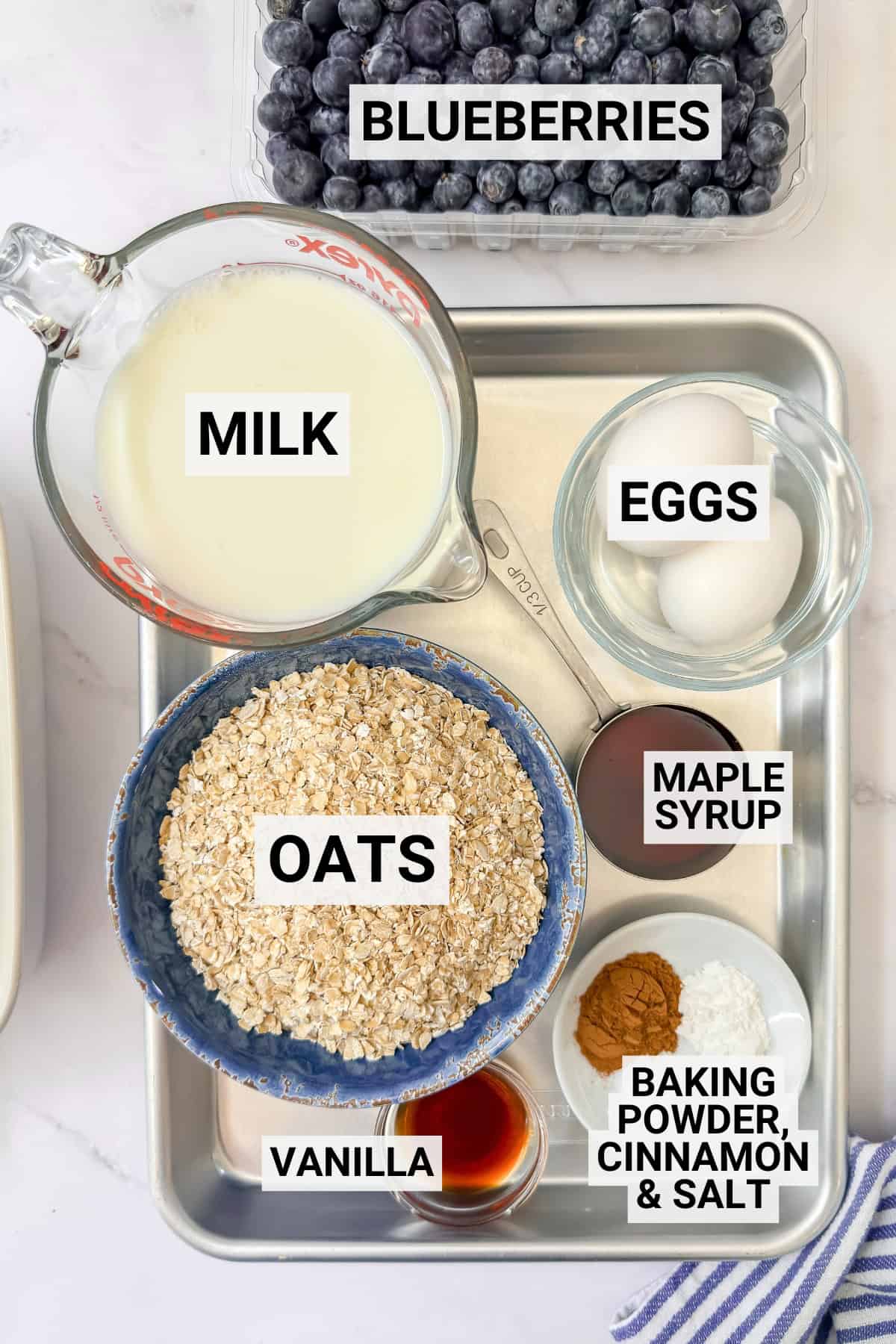 A tray with bowls and containers holding ingredients for blueberry Amish baked oatmeal: blueberries, milk, eggs, oats, maple syrup, vanilla, and a mix of baking powder, cinnamon, and salt-all neatly labeled.