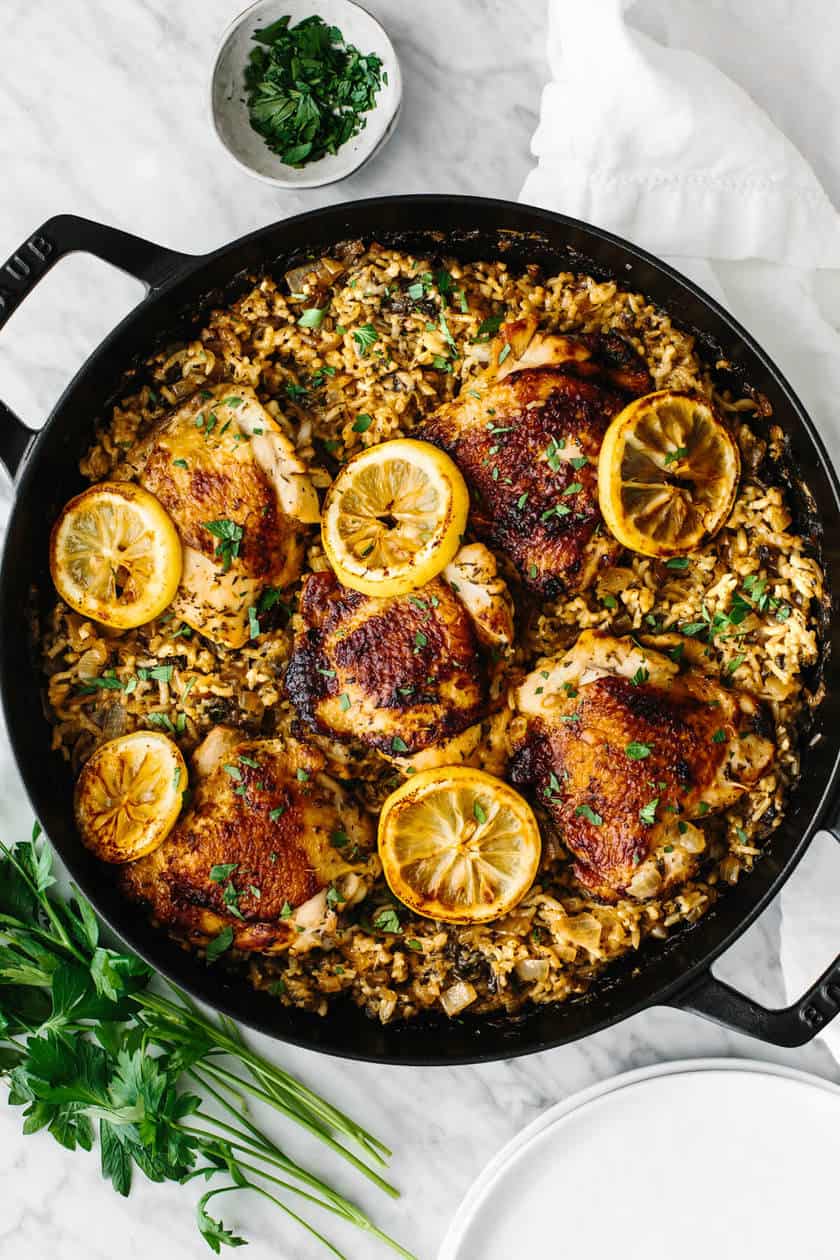 A skillet filled with baked chicken thighs on a bed of rice, garnished with lemon slices and chopped parsley, sits on a marble countertop-perfect for Mediterranean Diet Dinners.