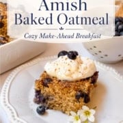 A slice of blueberry Amish baked oatmeal topped with whipped cream and fresh blueberries sits on a plate, accompanied by a cup of coffee, with the rest of the blueberry Amish baked oatmeal dish in the background.