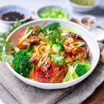 A bowl of Asian BBQ Chicken thighs glazed to perfection, served over noodles with broccoli, and garnished with sesame seeds, green onions, and cilantro. Soy sauce and seasonings are in the background.