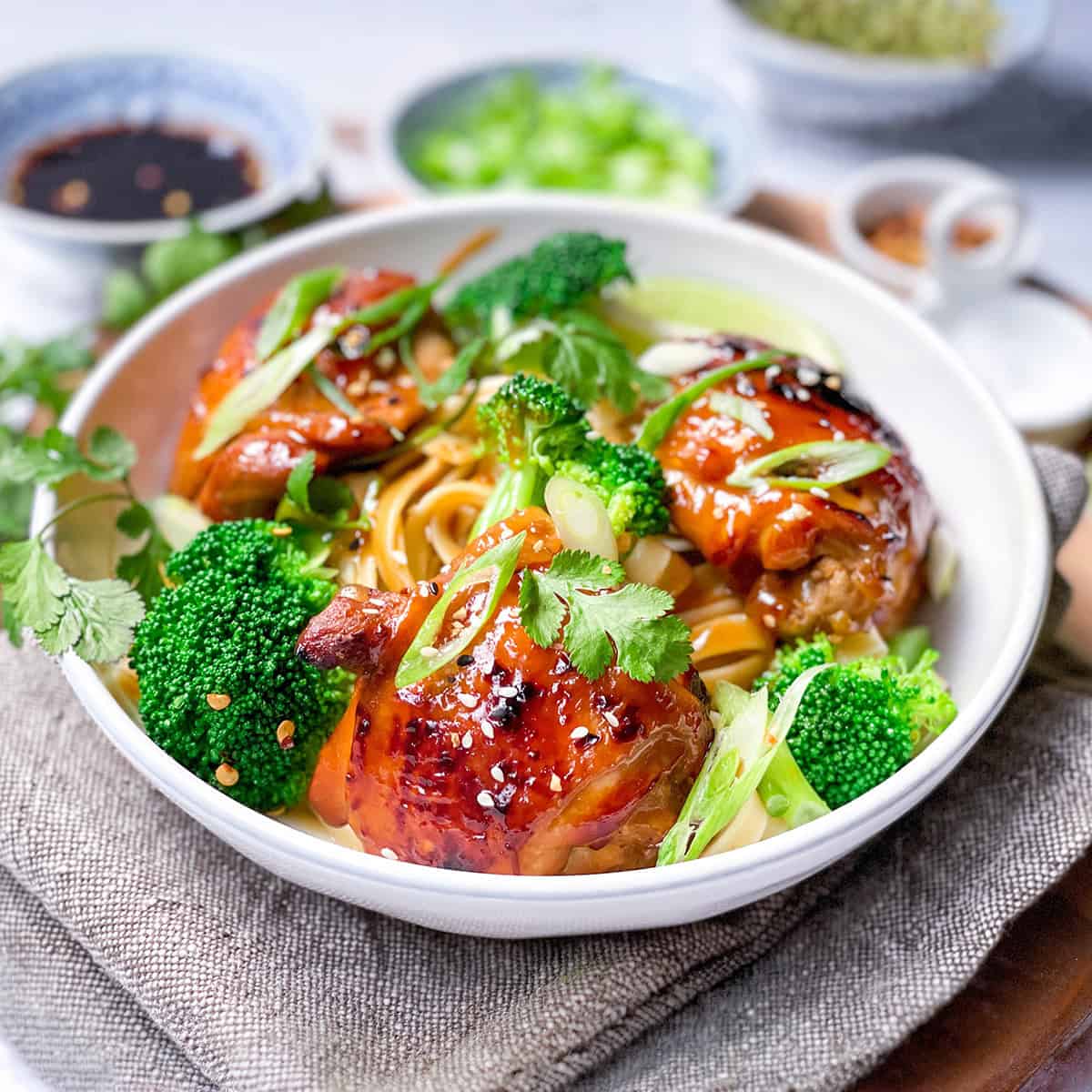 A bowl of Asian BBQ Chicken thighs glazed to perfection, served over noodles with broccoli, and garnished with sesame seeds, green onions, and cilantro. Soy sauce and seasonings are in the background.