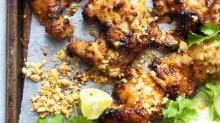 A healthy 4 ingredient dinners option: grilled chicken thighs with peanut sauce, chopped peanuts, lime wedges, cilantro, and sliced cucumber on a parchment-lined baking tray.