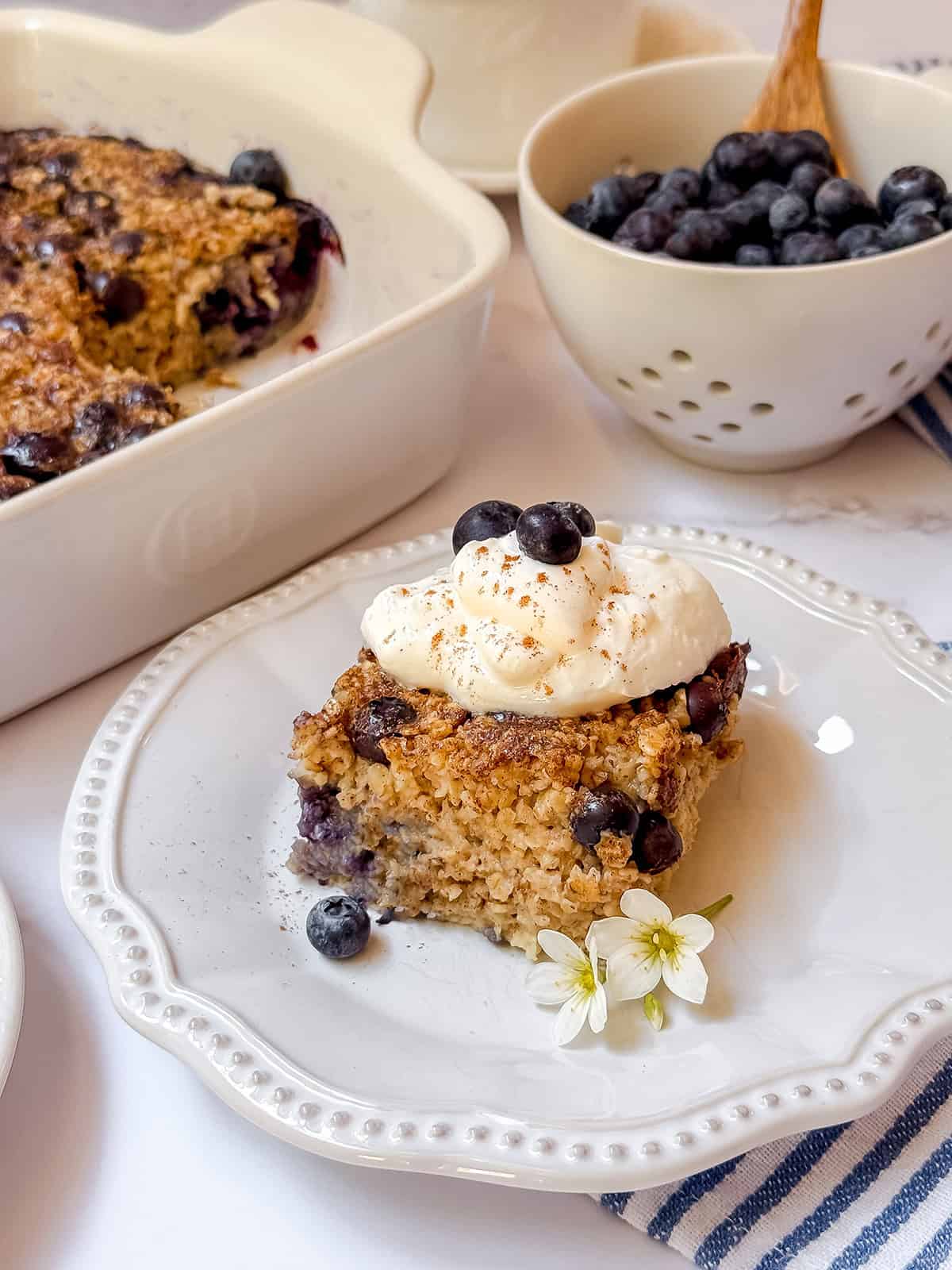 A slice of blueberry Amish baked oatmeal with blueberries, topped with whipped cream and extra berries on a white plate, with a baking dish and a bowl of blueberries in the background.