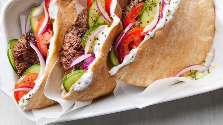 Two pita sandwiches filled with falafel, sliced cucumbers, tomatoes, and red onions, topped with white sauce—perfect for January Mediterranean Diet Dinners—served in a tray with a side of yogurt sauce.