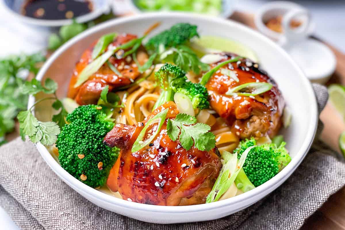 A bowl of Asian BBQ Chicken thighs glazed to perfection, served over noodles with broccoli, sliced green onions, cilantro, and sesame seeds.
