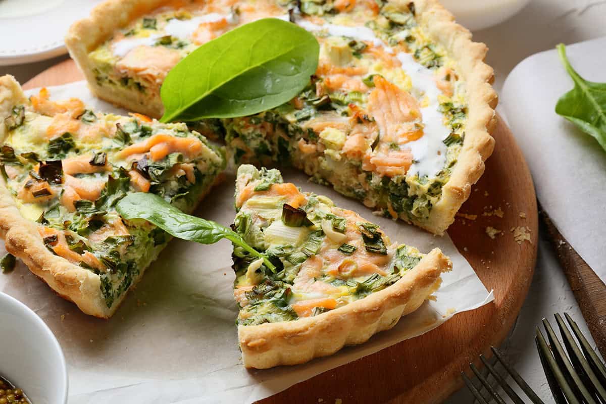 A sliced salmon quiche with spinach rests on a wooden board, topped with fresh spinach leaves as garnish.