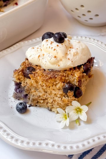 A square of blueberry Amish baked oatmeal topped with whipped cream sits on a white plate, garnished with a small white flower.