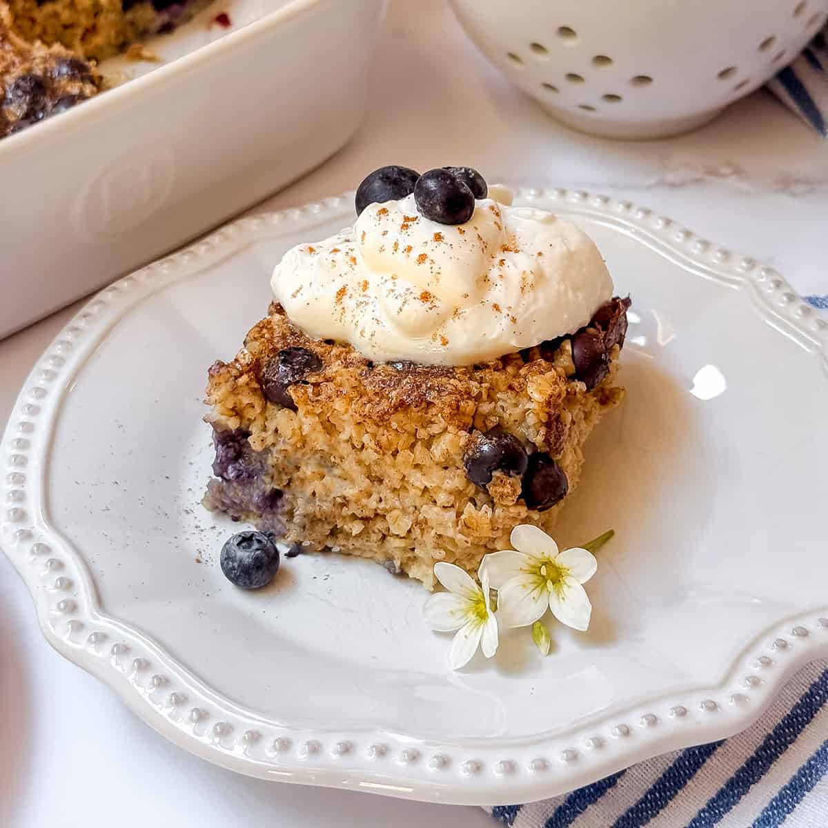 A square of blueberry Amish baked oatmeal topped with whipped cream sits on a white plate, garnished with a small white flower.