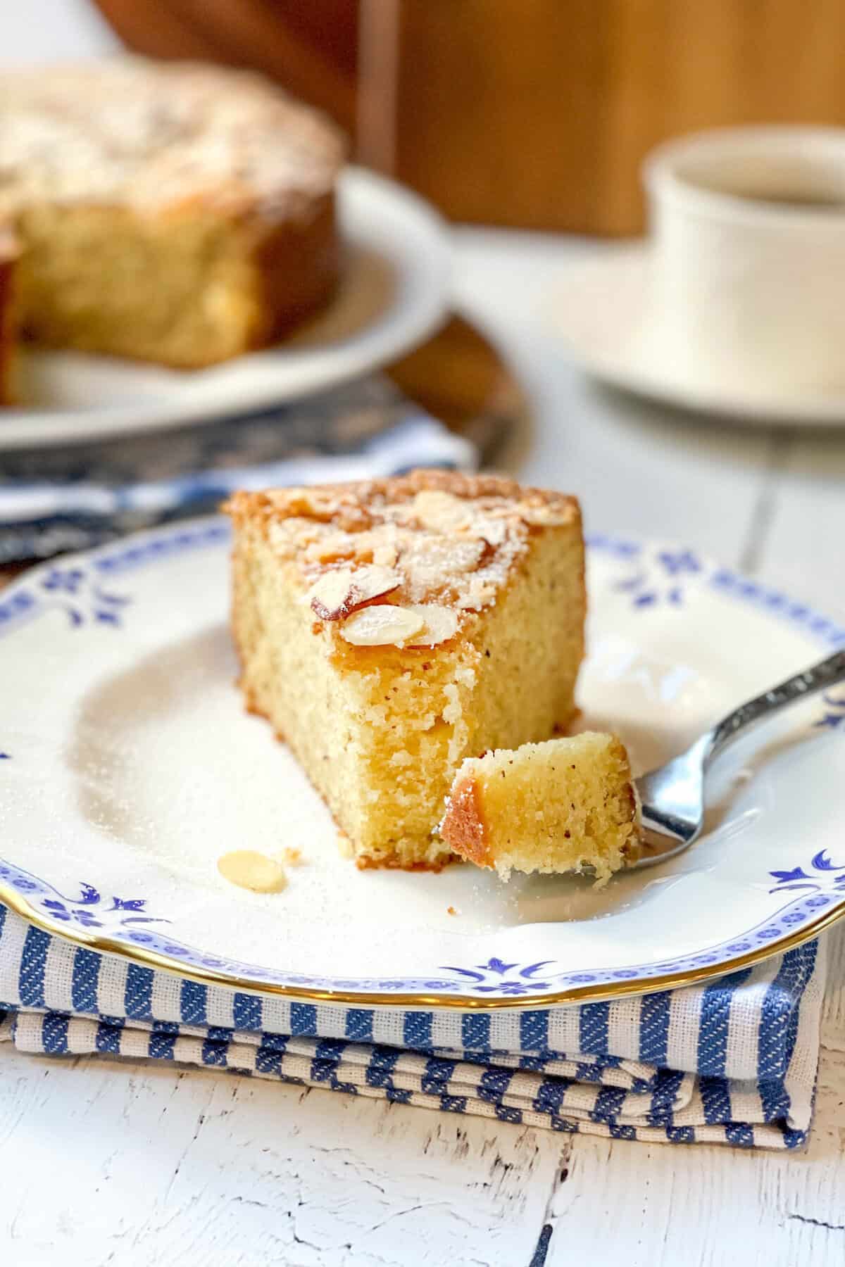 A delightful slice of almond cake with powdered sugar rests on a decorative plate, a fork holding a bite beside it, and a cup in the background.