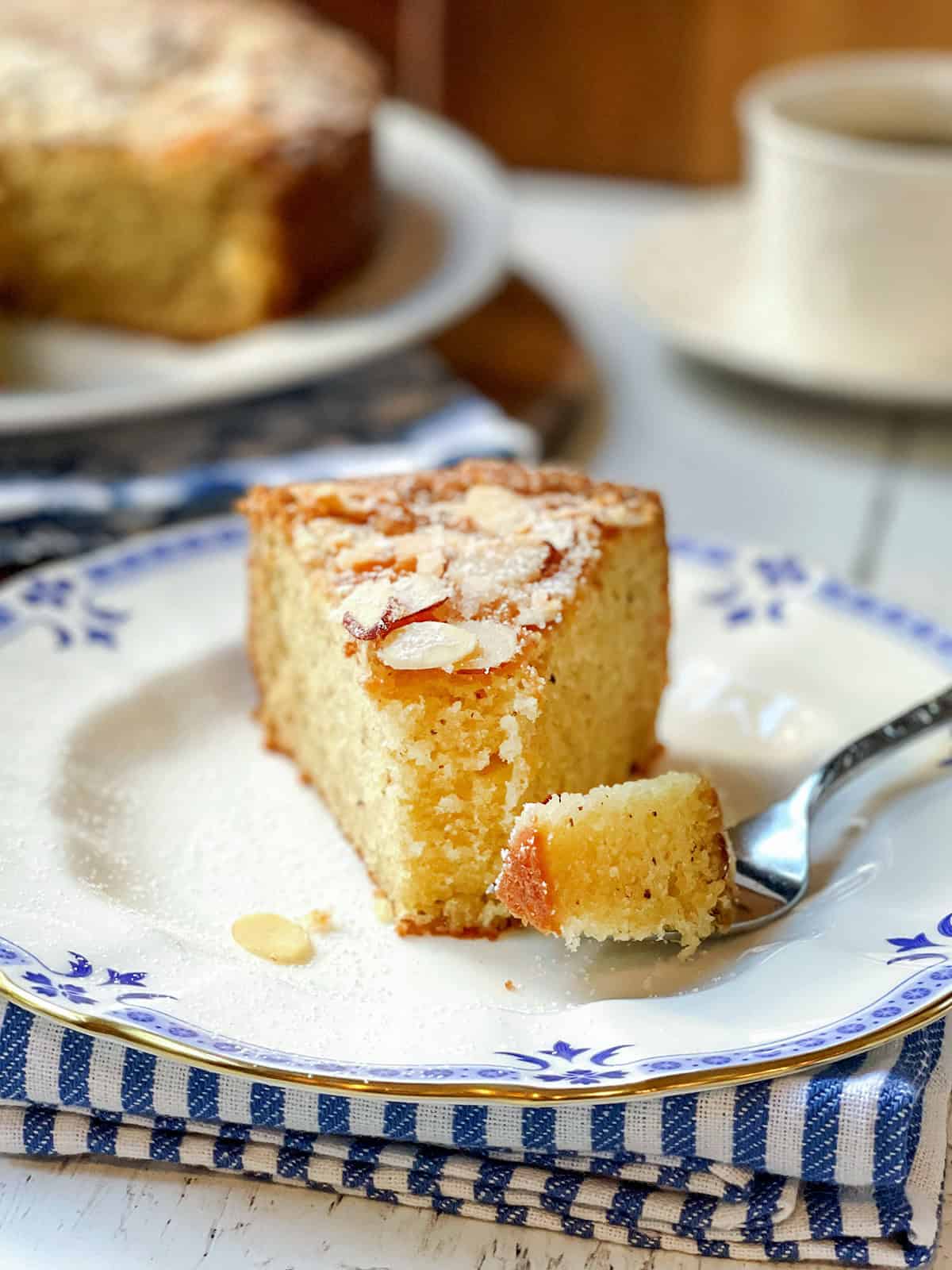 A slice of Almond Cake with powdered sugar and almond slices on top sits on a plate, with a fork holding a bite. In the background, another Almond Cake and a cup add to the inviting scene.