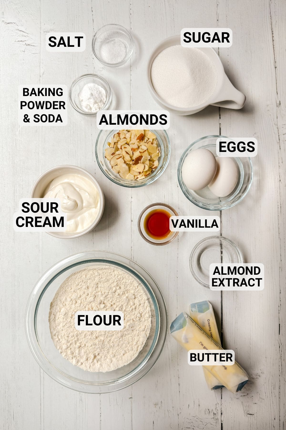 Various labeled baking ingredients for Almond Cake, including flour, butter, eggs, sugar, sour cream, sliced almonds, vanilla, almond extract, salt, and baking powder & soda arranged on a white surface.
