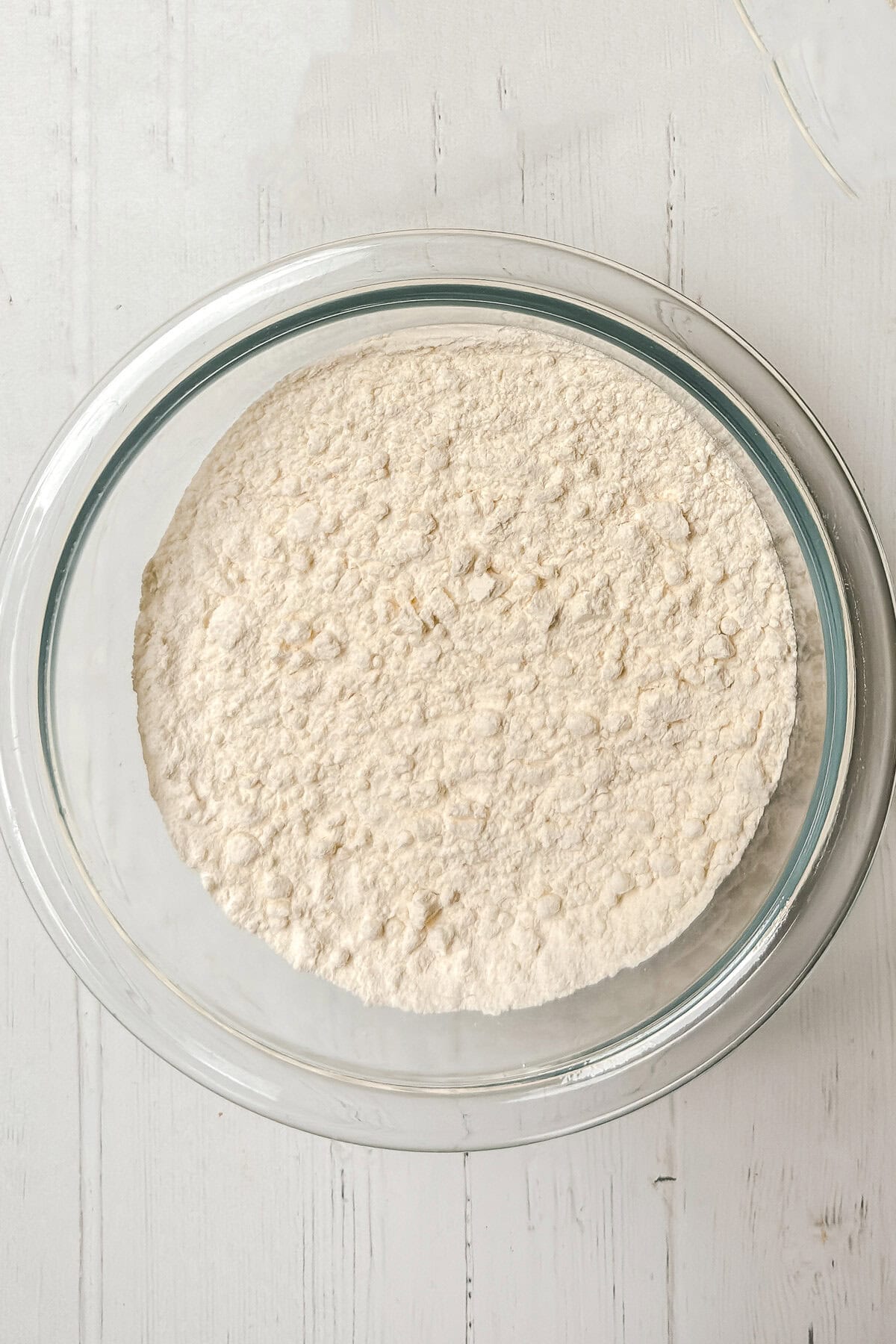 A glass bowl filled with almond cake flour mixture sits on a white wooden surface.