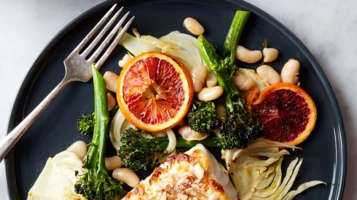 A plate with baked fish topped with sliced almonds, broccolini, white beans, fennel, and blood orange slices; a flavorful 30 Minute One Pot Dinner. Fork on the plate, bowl of oranges in the background.