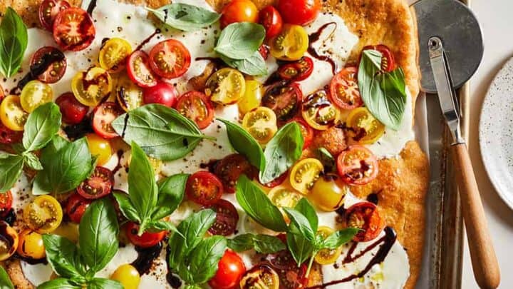 A rectangular flatbread topped with mozzarella cheese, red and yellow cherry tomato slices, fresh basil leaves, and a drizzle of balsamic glaze makes for one of the best Sheet Pan Mediterranean Diet Dinners—served next to a pizza cutter.