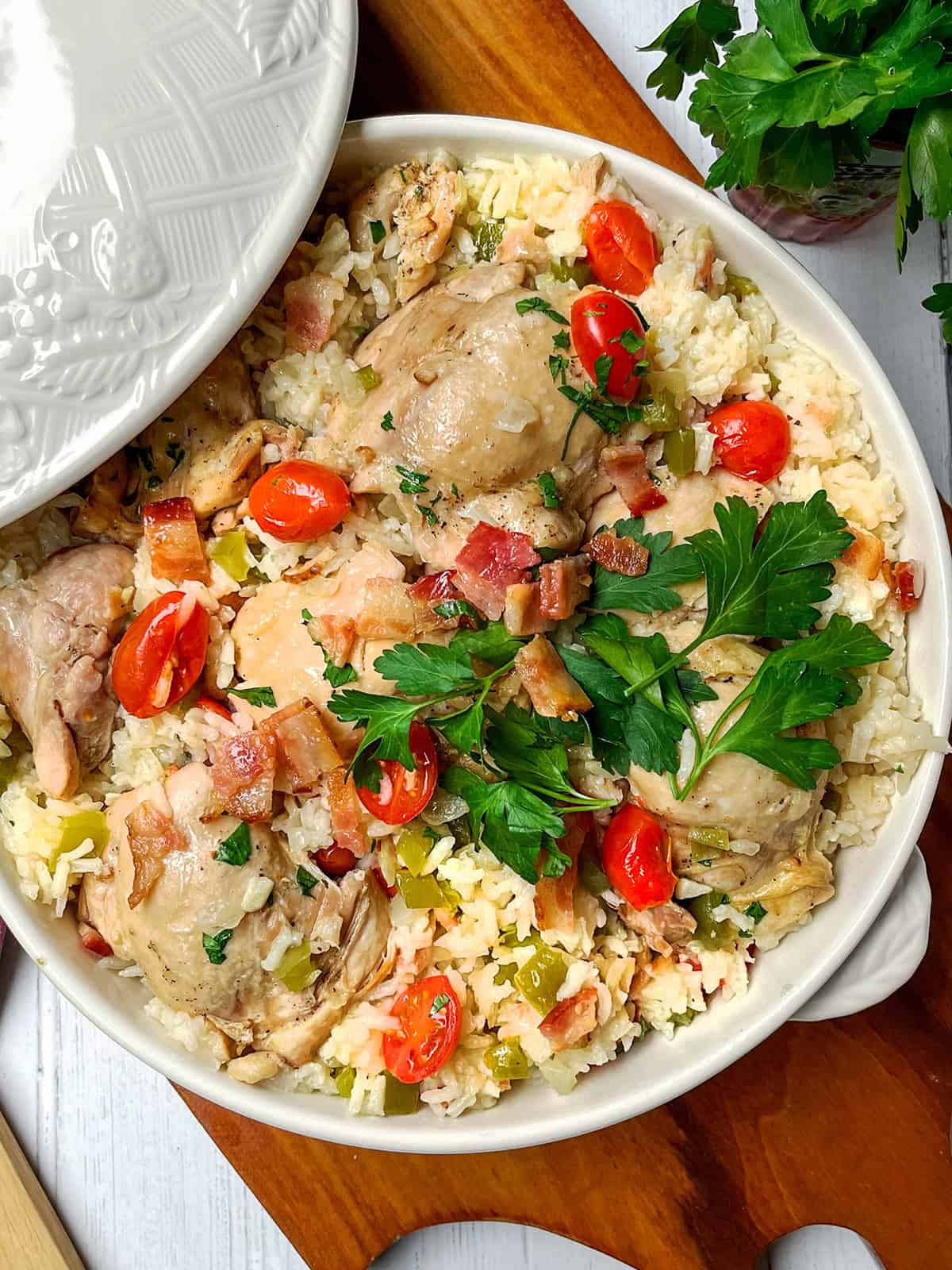 A casserole dish filled with Chicken Perloo—tender cooked chicken thighs, white rice, chopped bacon, cherry tomatoes, and parsley—rests on a wooden surface.