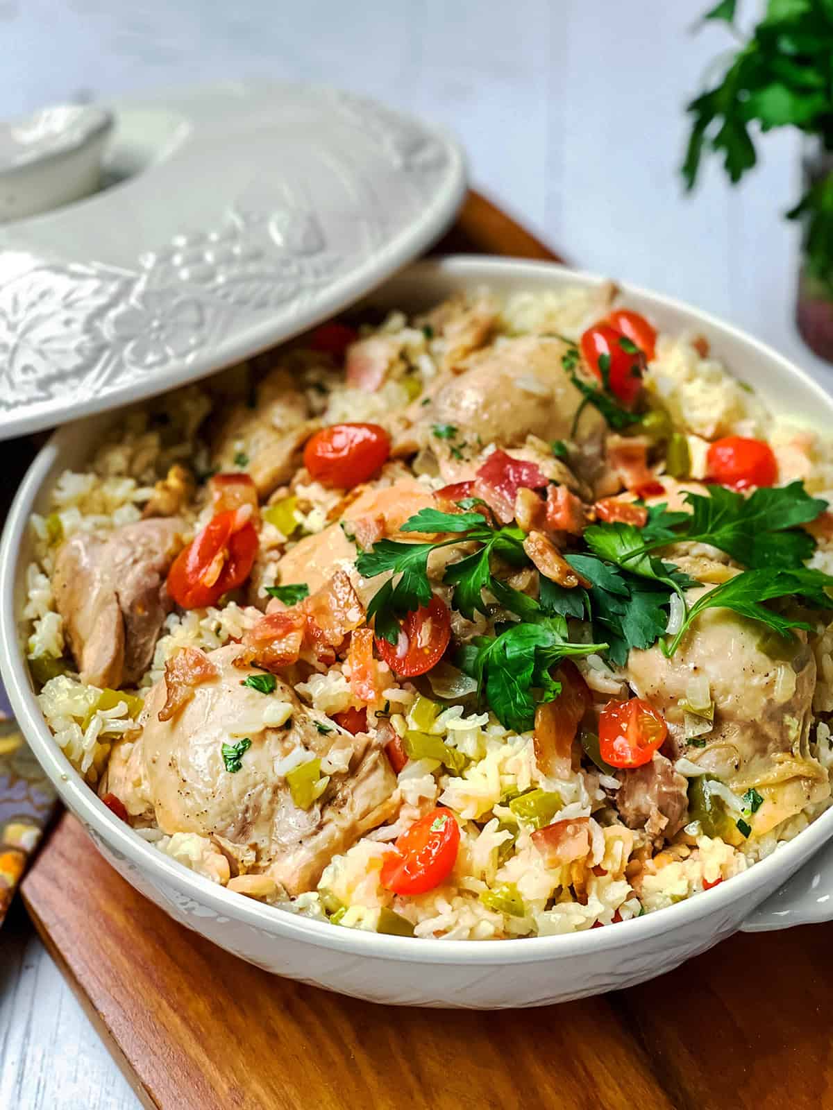 A ceramic dish filled with Chicken Perloo—cooked rice, chicken pieces, cherry tomatoes, and fresh parsley garnish—sits on a wooden board next to a partially removed lid.