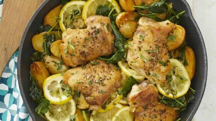 A skillet containing cooked chicken thighs, sliced lemons, potatoes, and leafy greens, with herbs sprinkled on top, sits on a blue and white patterned cloth—perfect for easy 30 Minute One Pot Dinners.