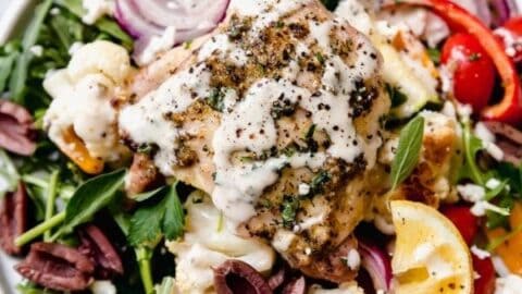 A plate of Mediterranean salad topped with grilled chicken, creamy dressing, cherry tomatoes, olives, red onion, cucumber, cauliflower, and lemon wedges—perfect for those who love easy Sheet Pan Mediterranean Diet Dinners.