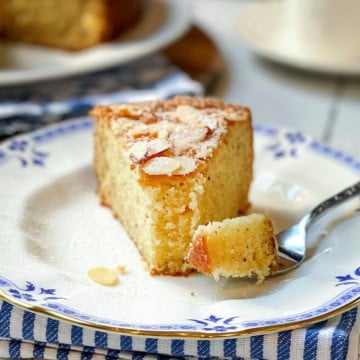 A slice of Almond Cake with sliced almonds on top sits on a decorative plate, while a fork holds a tender piece of the cake.