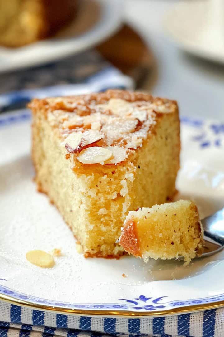 A slice of Almond Cake with sliced almonds on top sits on a decorative plate, while a fork holds a tender piece of the cake.