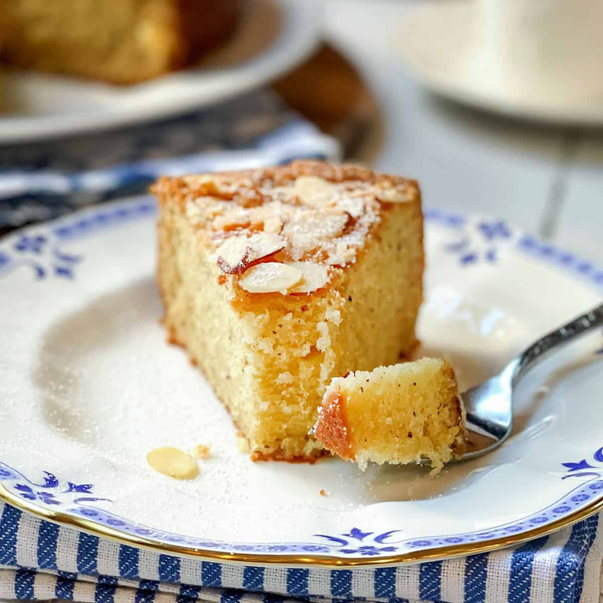A slice of Almond Cake with sliced almonds on top sits on a decorative plate, while a fork holds a tender piece of the cake.