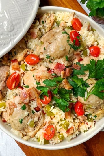 A ceramic dish filled with Chicken Perloo—rice, chicken pieces, chopped bacon, cherry tomatoes, and fresh parsley—with a wooden spoon and parsley on the side.