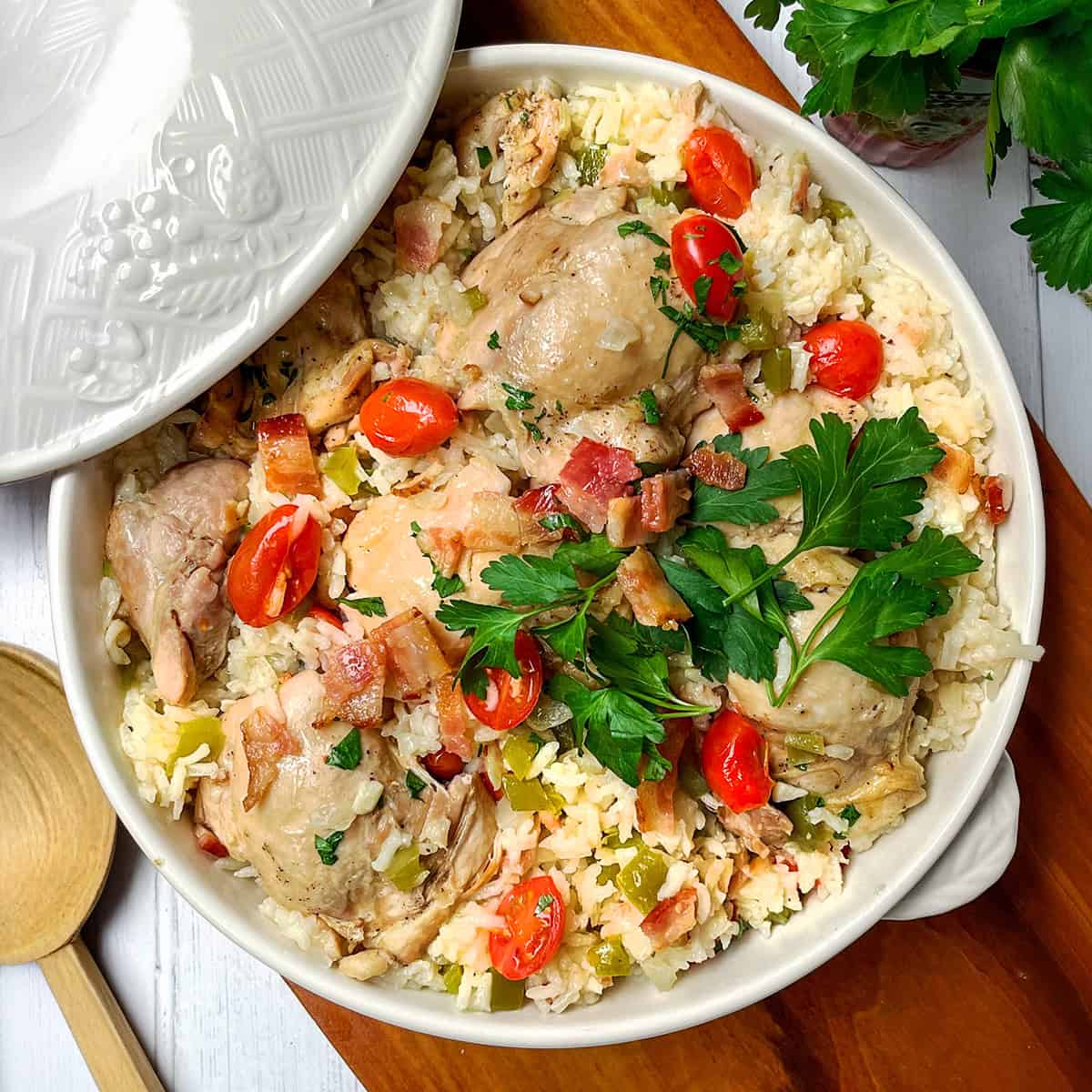 A ceramic dish filled with Chicken Perloo—rice, chicken pieces, chopped bacon, cherry tomatoes, and fresh parsley—with a wooden spoon and parsley on the side.