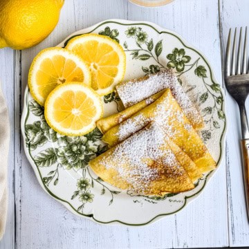 Three folded crepes, perfect for Shrove Tuesday Pancakes, are dusted with powdered sugar and paired with three lemon slices on a floral-patterned plate, with a fork and a lemon nearby on a white wooden surface.