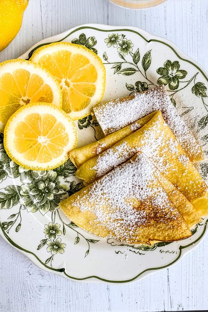 Three folded crepes, perfect for Shrove Tuesday Pancakes, are dusted with powdered sugar and paired with three lemon slices on a floral-patterned plate, with a fork and a lemon nearby on a white wooden surface.
