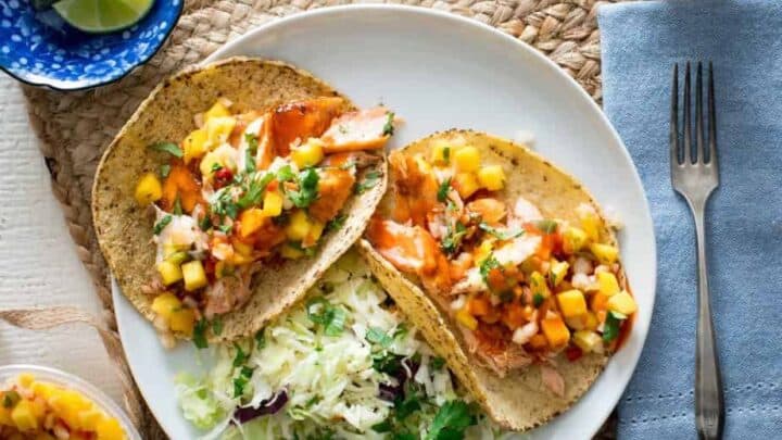 Two fish tacos topped with mango salsa and chopped cilantro are served on a white plate with a side of slaw—perfect for quick 20 Minute Mediterranean Dinners. Lime wedges and hot sauce are nearby.