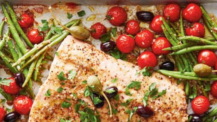 Baked salmon fillet on a tray with green beans, cherry tomatoes, olives, and caper berries is an ideal choice for Sheet Pan Mediterranean Diet Dinners, garnished with herbs and served with mustard sauce beside a striped cloth.