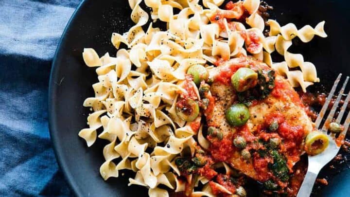 A black plate with egg noodles and chicken in tomato sauce with olives and herbs, next to a small bowl of ground black pepper—a perfect choice for quick 20 Minute Mediterranean Dinners.