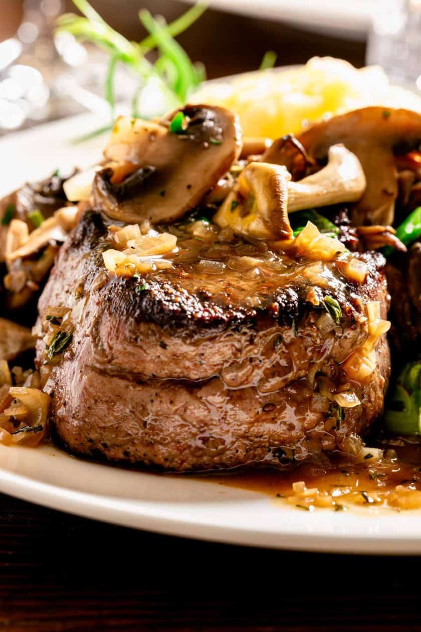 Close-up of a juicy steak topped with saut&eacute;ed mushrooms and onions, garnished with fresh herbs-the perfect centerpiece for romantic dinner recipes. Cooked to perfection and served on a white plate, it highlights the rich colors and textures of this intimate dining experience.