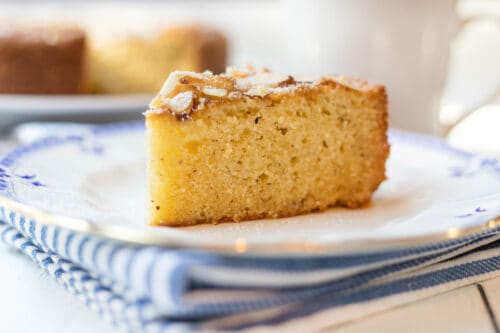 A slice of almond cake topped with powdered sugar sits on a white plate with a blue rim, placed on a folded blue and white striped napkin.