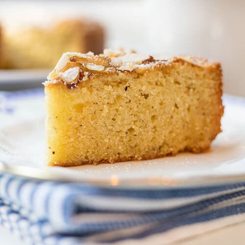 A slice of almond cake topped with powdered sugar sits on a white plate with a blue rim, placed on a folded blue and white striped napkin.