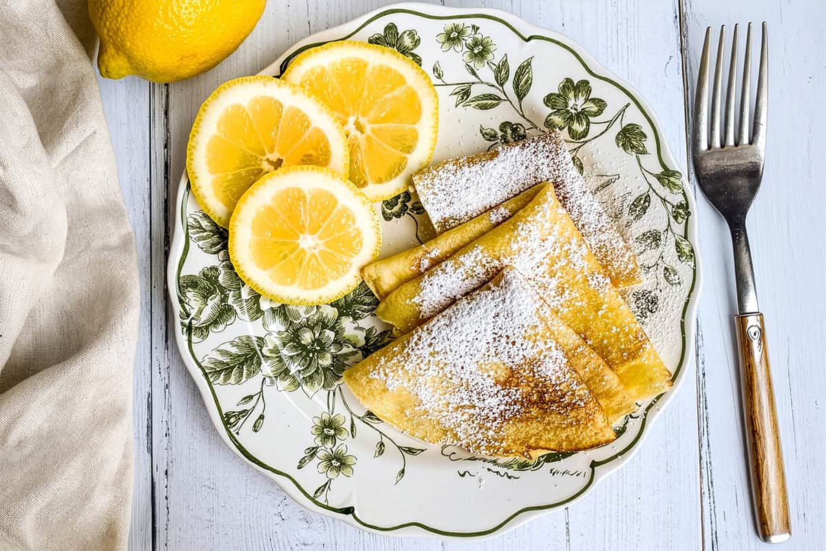 A decorative plate with Shrove Tuesday Pancakes-folded crepes dusted with powdered sugar, garnished with lemon slices, a whole lemon, and a fork on a white wooden table.