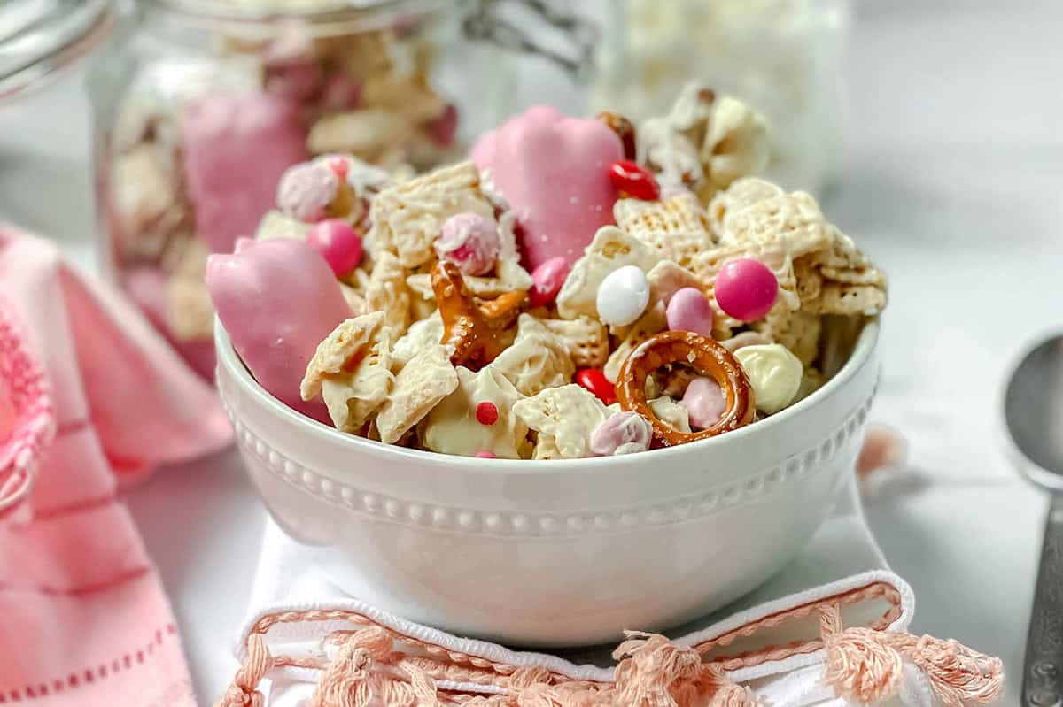 A white bowl filled with Valentine's Day Crack snack mix-pink and white coated pretzels, cereal pieces, and colorful candy-sits on a napkin with a pink ribbon.