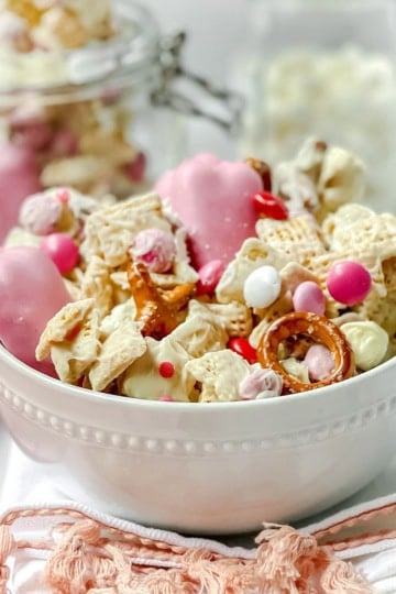 A white bowl filled with Valentine's Day Crack snack mix—cereal, pretzels, pink and white candy-coated chocolates, and white chocolate-dipped pieces. In the background, a jar holds even more of this festive treat.
