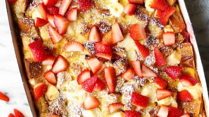 A rectangular baking dish filled with bread pudding topped with sliced strawberries and a dusting of powdered sugar, perfect for Spring Brunch Recipes, surrounded by plates and extra strawberries on a marble surface.
