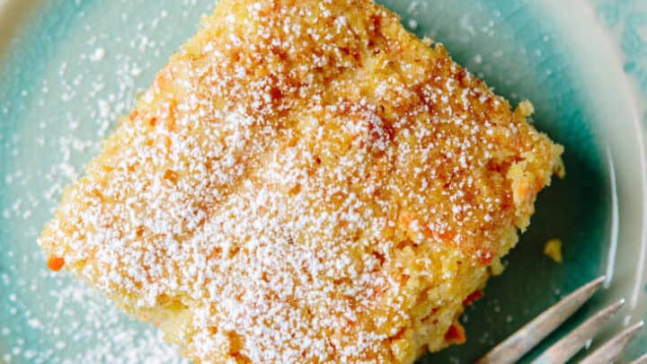 A square piece of cake topped with powdered sugar sits on a blue-green plate next to a fork, making a delightful finish to your March Mediterranean Diet Dinners.
