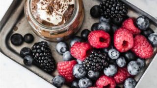 A metal tray with a jar of chocolate mousse topped with whipped cream and chocolate shavings, next to fresh raspberries, blackberries, and blueberries—perfect for adding a sweet finish to your April Clean Eating Dinners.